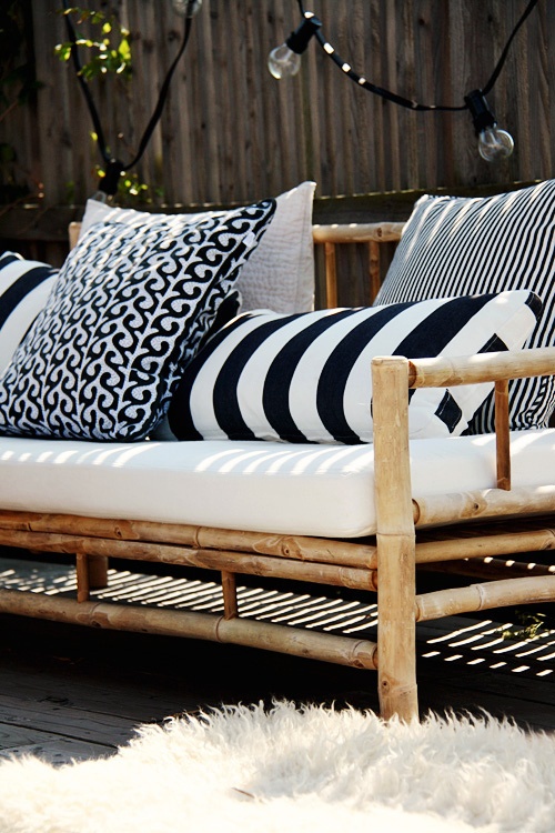 A bright outdoor lounge area with a bamboo daybed dressed in black-and-white patterned pillows, white cushion, and string lights overhead, framed by lush greenery and a wooden fence.