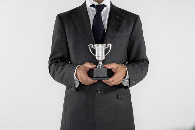A person in a dark suit holding a small trophy against a neutral gray background, conveying achievement and professionalism